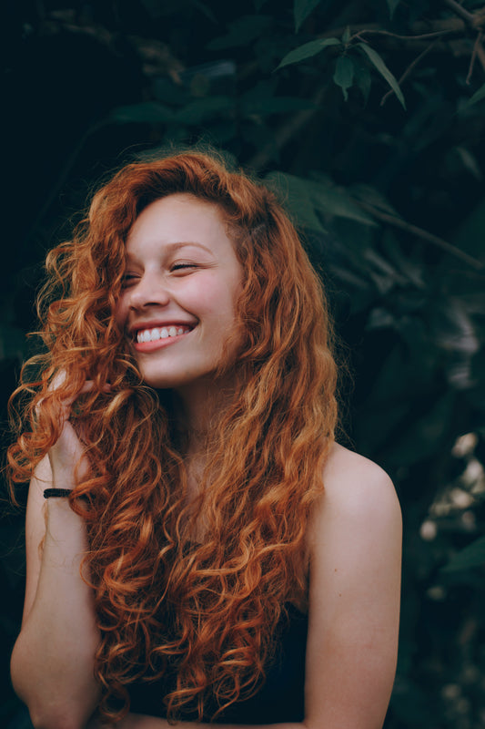Girl with curly red hair smiles and looks off into the distance, as the image for a blog post on preventing hair flyaways